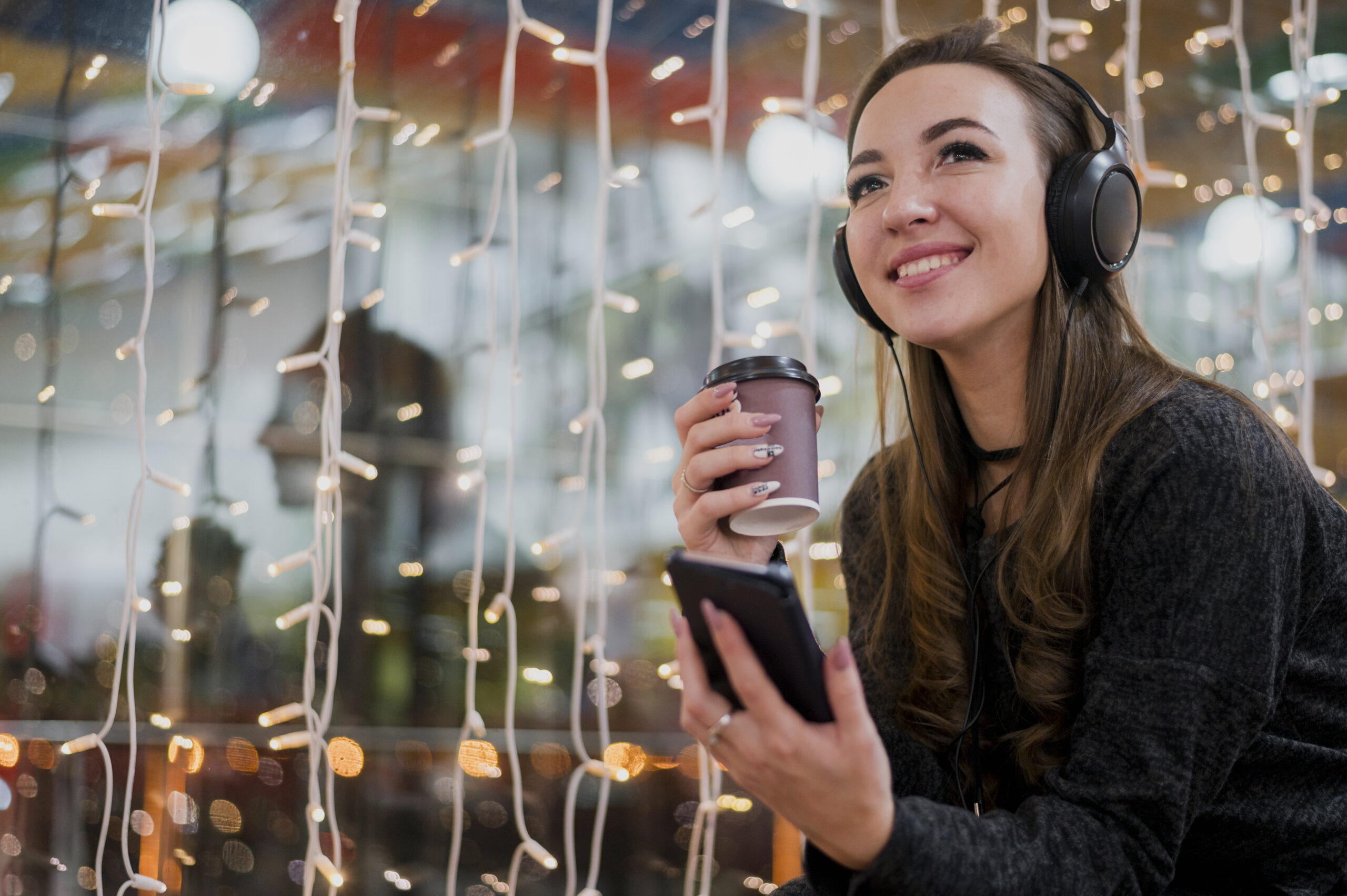 portrait-smiling-woman-wearing-headphones-holding-cup-phone-near-christmas-lights-scaled-1